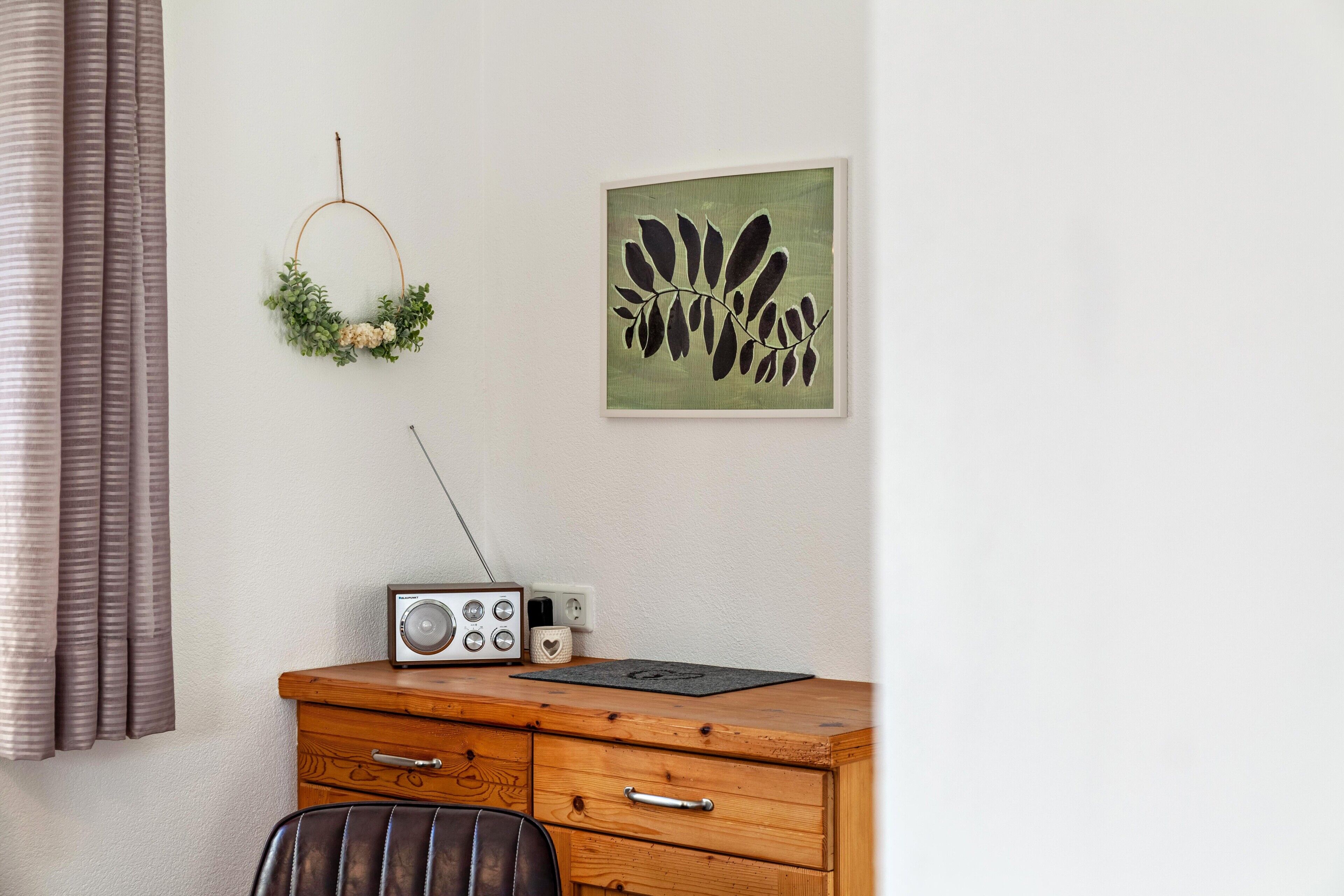 Une pièce comprenant une commode en bois, une radio, une couronne décorative accrochée au mur et une œuvre d’art encadrée.