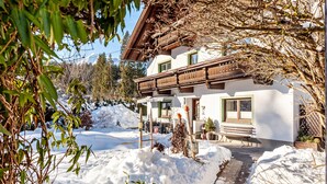 A two-story chalet with a wooden balcony, a snow-covered yard, and a bench outside. - Apartment "Alpenrose" with Mountain View, Shared Terrace & Wi-Fi (Leutasch)