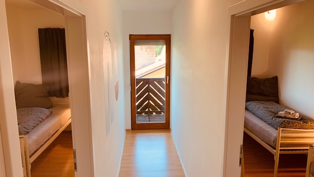 Un estrecho pasillo con dos camas individuales a cada lado, suelo de madera y una puerta con diseño geométrico.