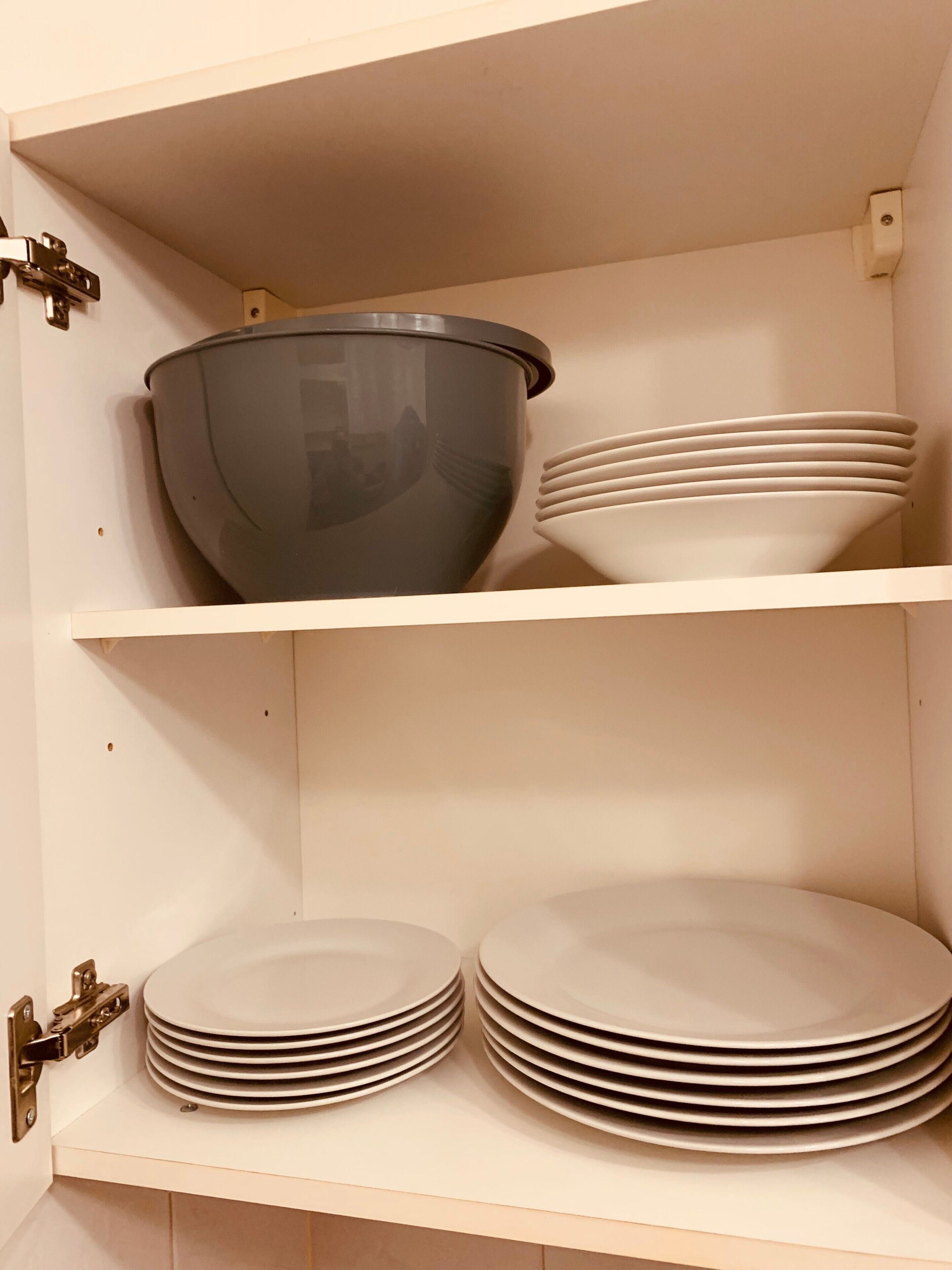 Ein Küchenschrank mit einer großen, dunkel gefärbten Rührschüssel auf der oberen Ablage und mehreren gestapelten weißen Tellern auf der unteren Ablage.