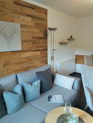 Een moderne woonkamer met een grijze bank, decoratieve kussens, een houten wandpaneel, een vloerlamp en een klein tafeltje met een glazen blad.