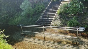 Une rambarde métallique surplombe une zone recouverte de petits graviers, avec de la verdure et un escalier en arrière-plan.