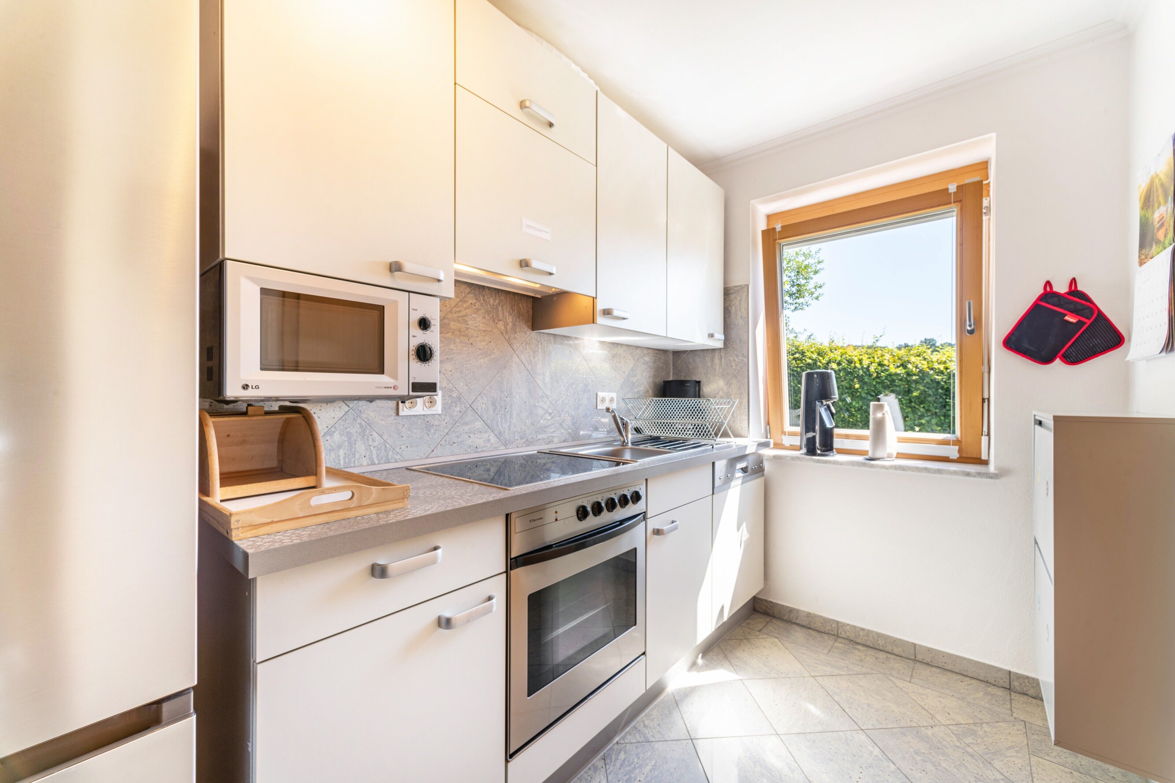 A modern kitchen with white cabinets, a built-in microwave, a stove, and a window with a view of greenery.