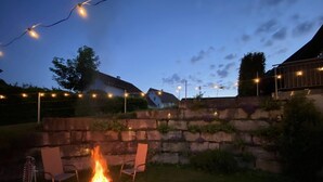Un muret en pierre avec un foyer et des chaises, entouré d’une pelouse et illuminé de guirlandes lumineuses.