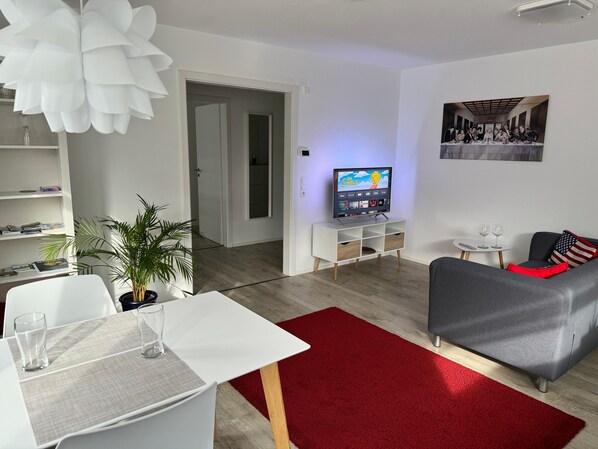Un salon moderne avec un téléviseur à écran plat, une table à manger avec des chaises, un tapis rouge et un canapé gris.