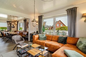 A modern living room with a leather sofa, dining table, and chairs, large windows with curtains, and a view of a house.