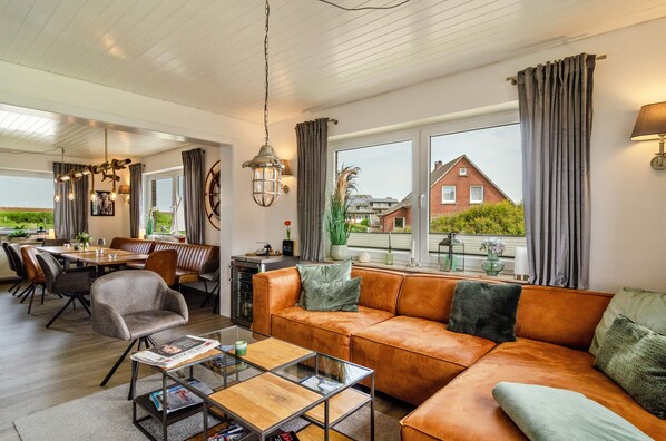 A modern living room with a leather sofa, dining table, and chairs, large windows with curtains, and a view of a house.
