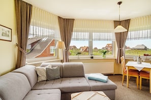 A living room with a sofa, a dining table, and a view of houses through large windows.