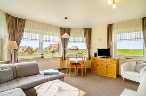 A room with a sofa, dining table, TV, and a view of houses through large windows.