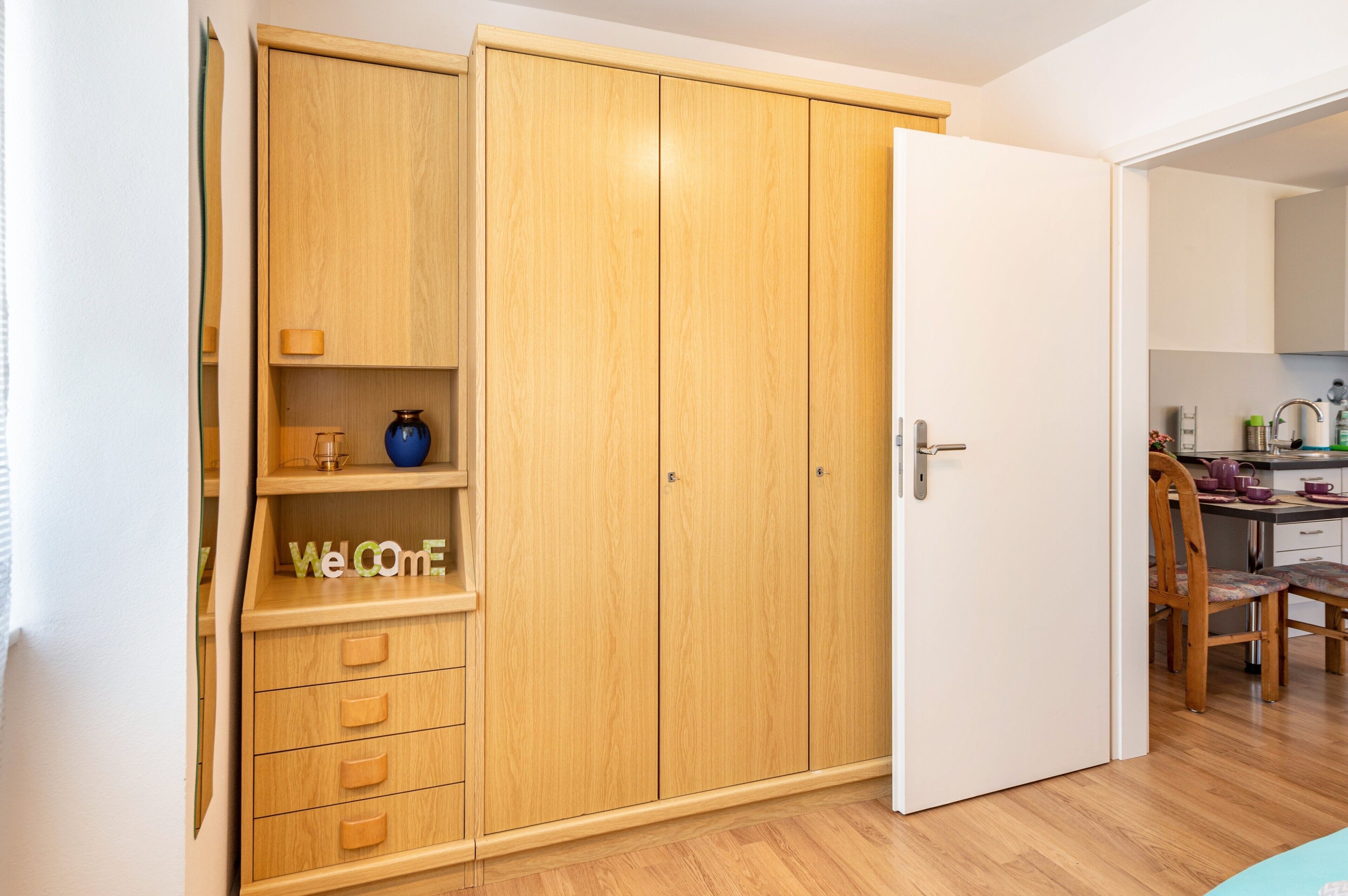 Une pièce avec une armoire en bois, une porte blanche et un coin cuisine comprenant une chaise et une table.