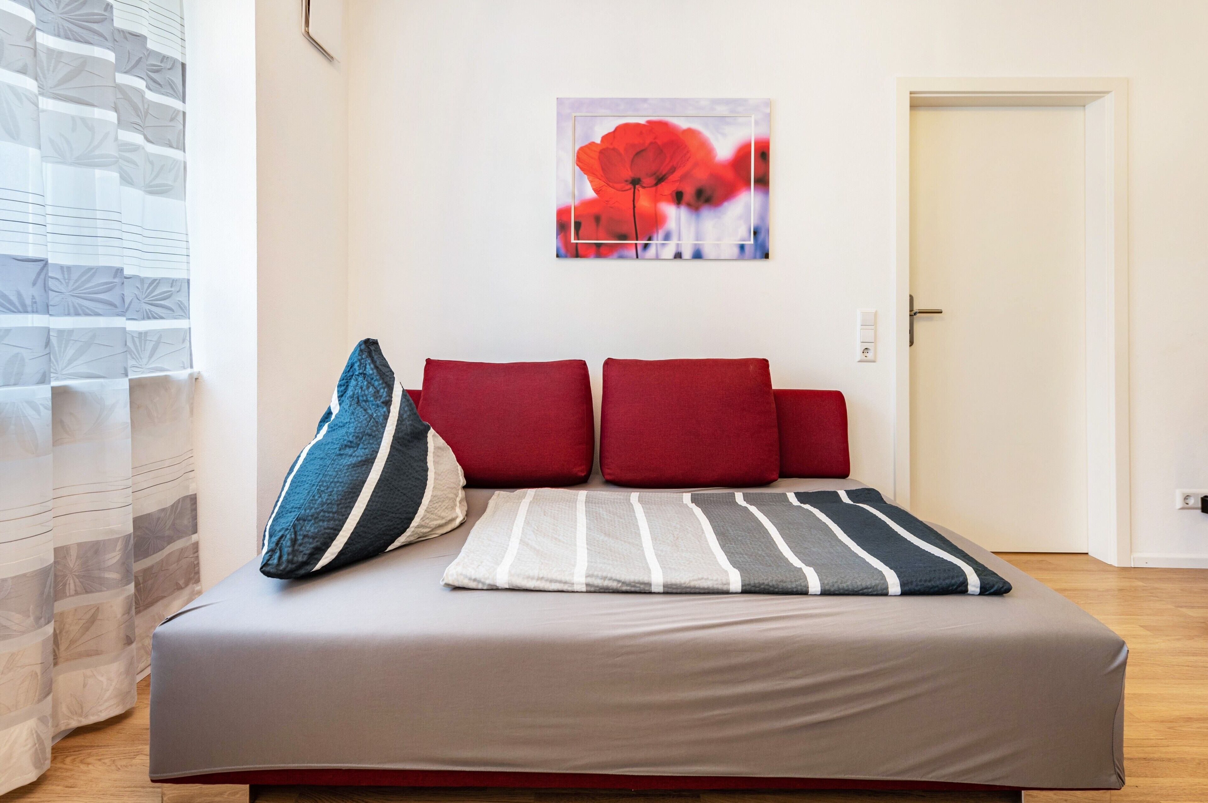 Una cama con una colcha a rayas grises y blancas, almohadas rojas y azules, y un cuadro de flores rojas en la pared.