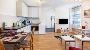 A modern kitchen with white cabinets, a dining table with chairs, a TV mounted on the wall, and a small seating area with a red chair and a plant.