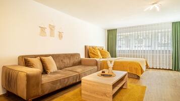 A modern hotel room with a sofa, a bed with yellow bedding, a wooden coffee table, and a large window with sheer curtains.