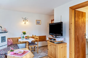 Een kamer met een houten kast, een televisie erboven, een eettafel met stoelen, een bankje en een wandlamp.