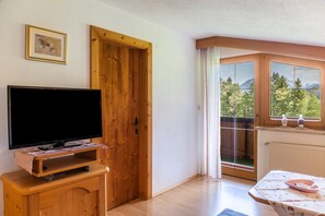 Ein Zimmer mit Fernseher auf einem Holztisch, einer Tür, einem Fenster mit Blick auf Bäume und Berge sowie einem Tisch mit Blumenmuster-Tischdecke.