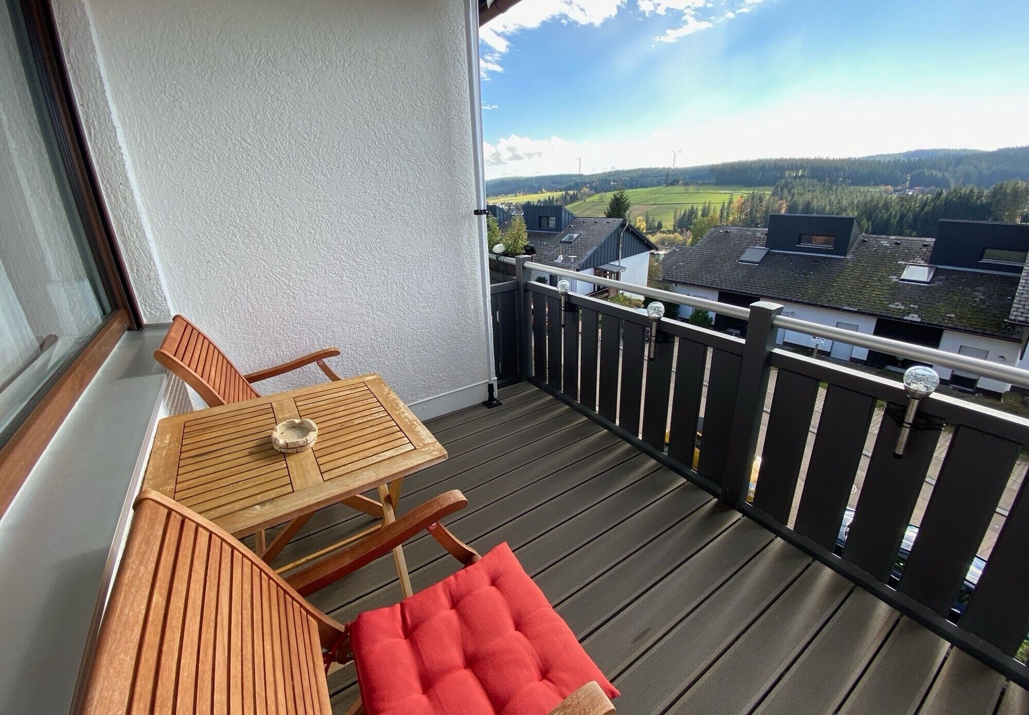 Un balcon avec une table et des chaises en bois, un coussin rouge et une vue sur un paysage verdoyant parsemé de bâtiments.