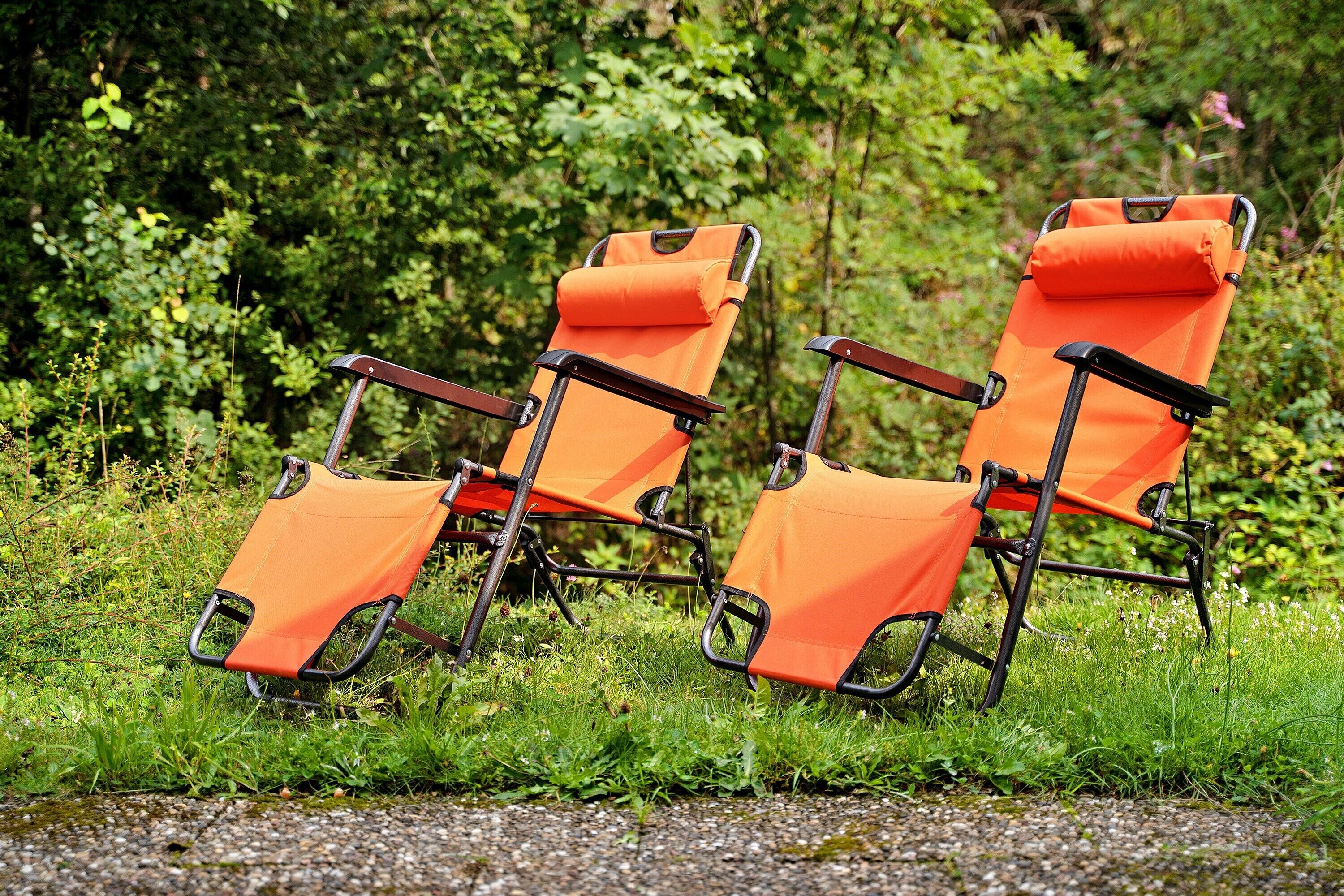 Two orange folding chairs with metal frames, positioned on a gravel path with greenery in the background.