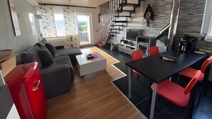 A modern living room with a sofa, coffee table, and red chairs. A staircase leads to a kitchen area with a TV and a fireplace. - Apartment 'Klug' with Mountain View, Private Terrace and Wi-Fi (Schonach im Schwarzwald)
