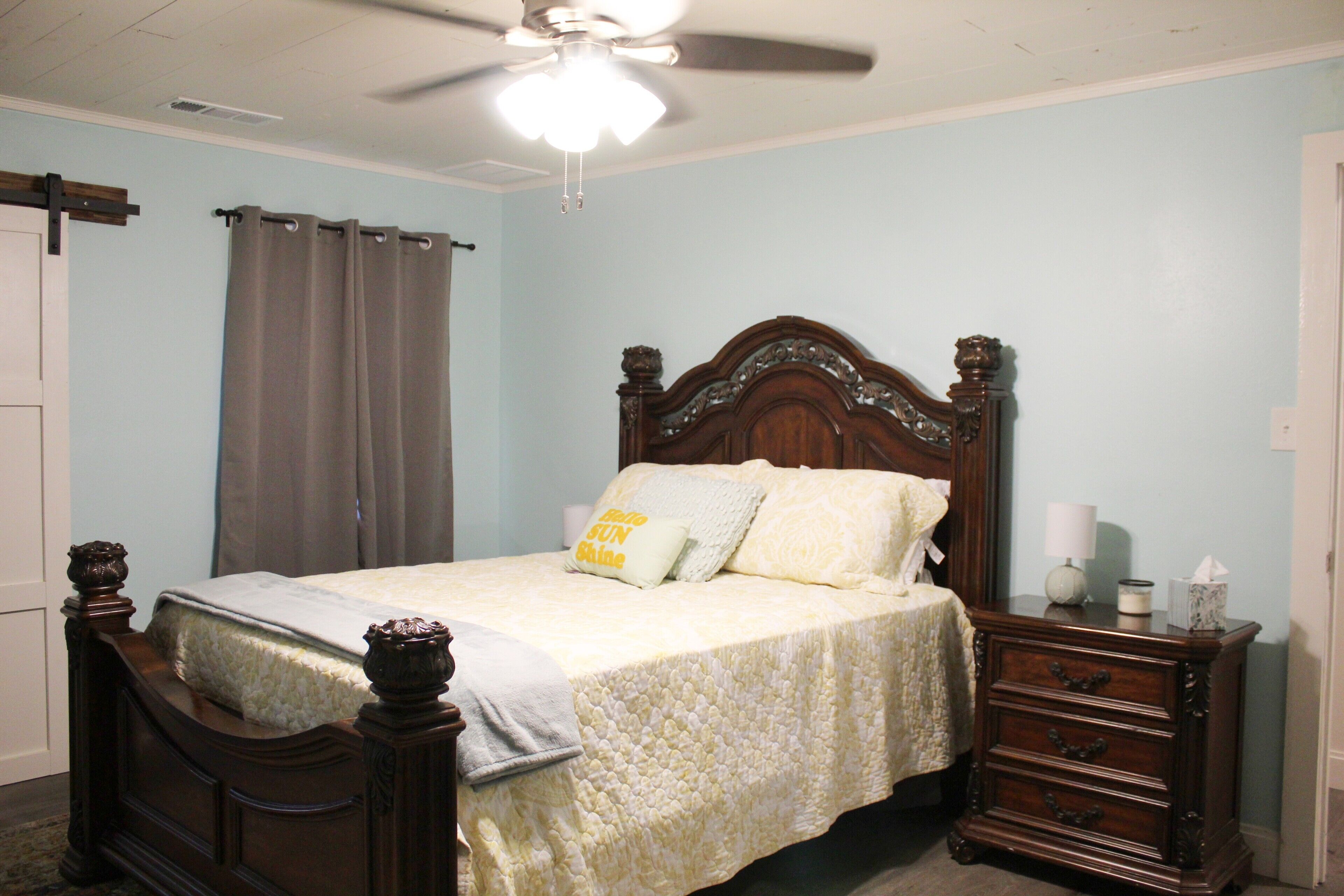 A bedroom with a wooden bed, a nightstand, a ceiling fan, and a window with curtains.