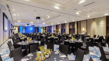 A large banquet hall with round tables, black chairs, and a stage with a large screen.