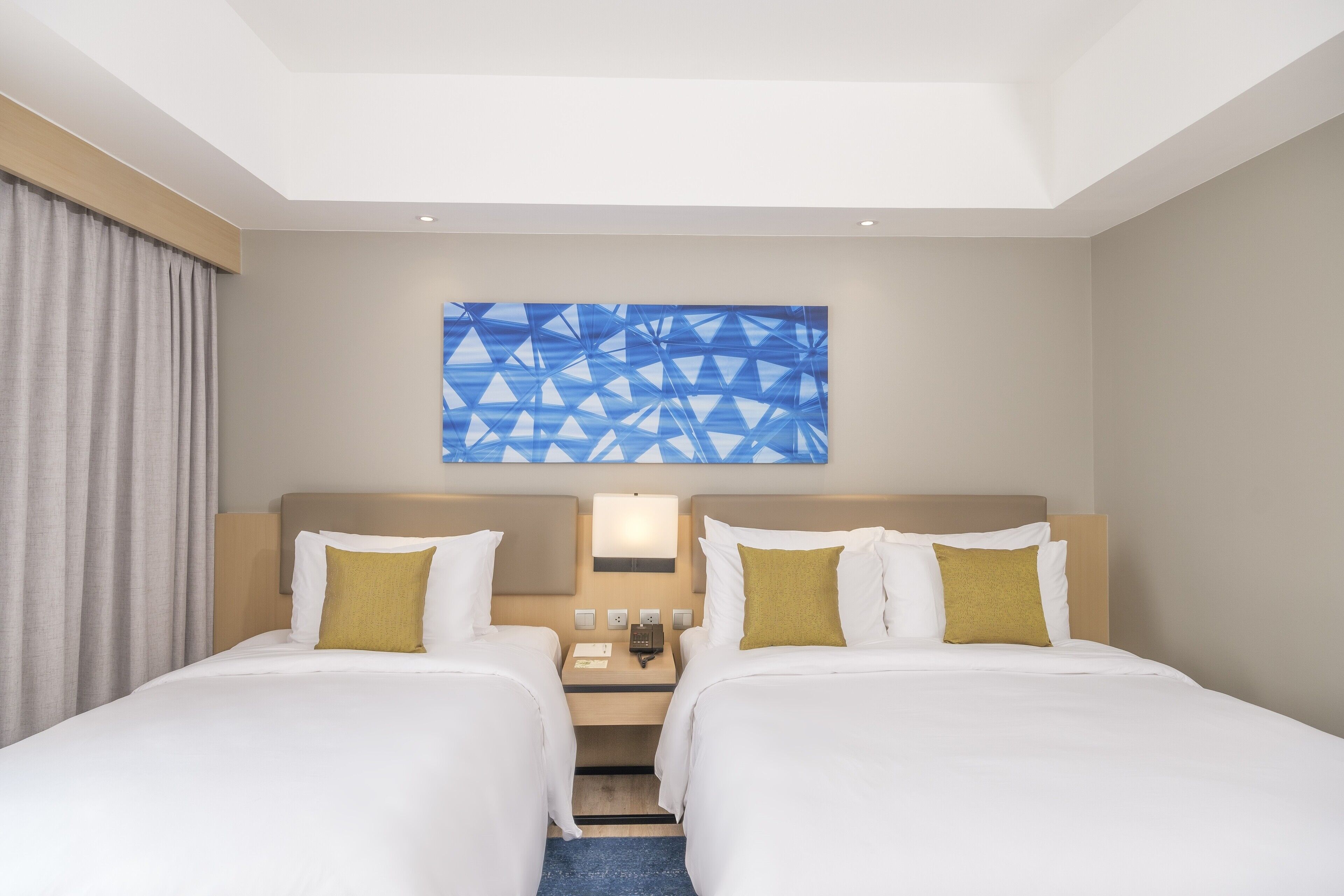 A hotel room with two beds, a nightstand, a wall-mounted lamp, and a geometric artwork above the beds.