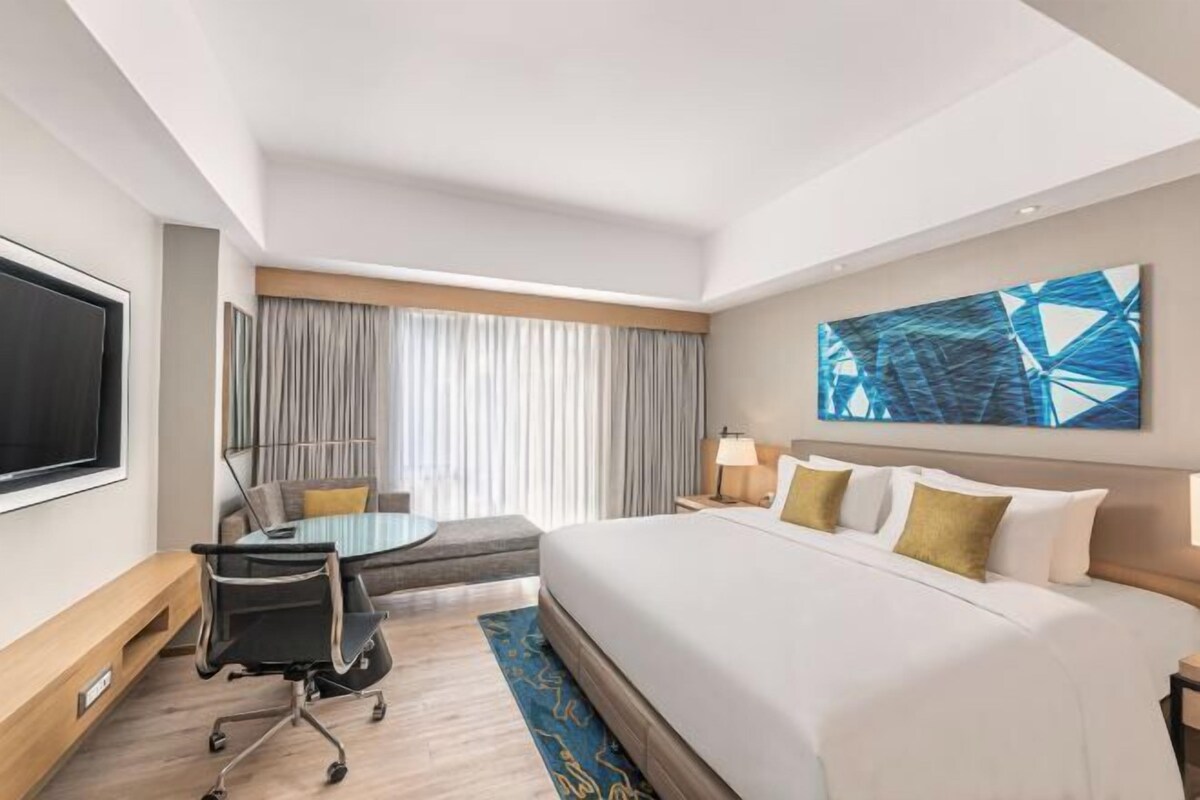 A hotel room with a large bed, a desk with a chair, a TV, and a painting on the wall.