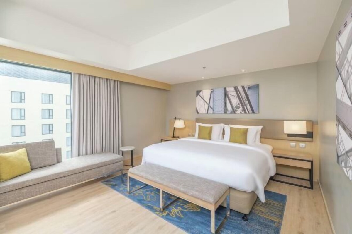 A modern hotel room with a large bed, a bench, a sofa, and a view of buildings through the window.