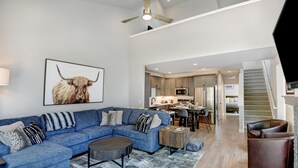 Un salon moderne avec un canapé d’angle bleu, une table basse et un tapis. On aperçoit, à travers une porte ouverte, un coin cuisine avec une table à manger et des chaises.