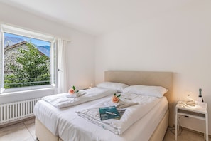 Habitación de hotel con cama, mesita de noche con una botella y vasos, y ventana con vistas a zonas verdes y montañas.