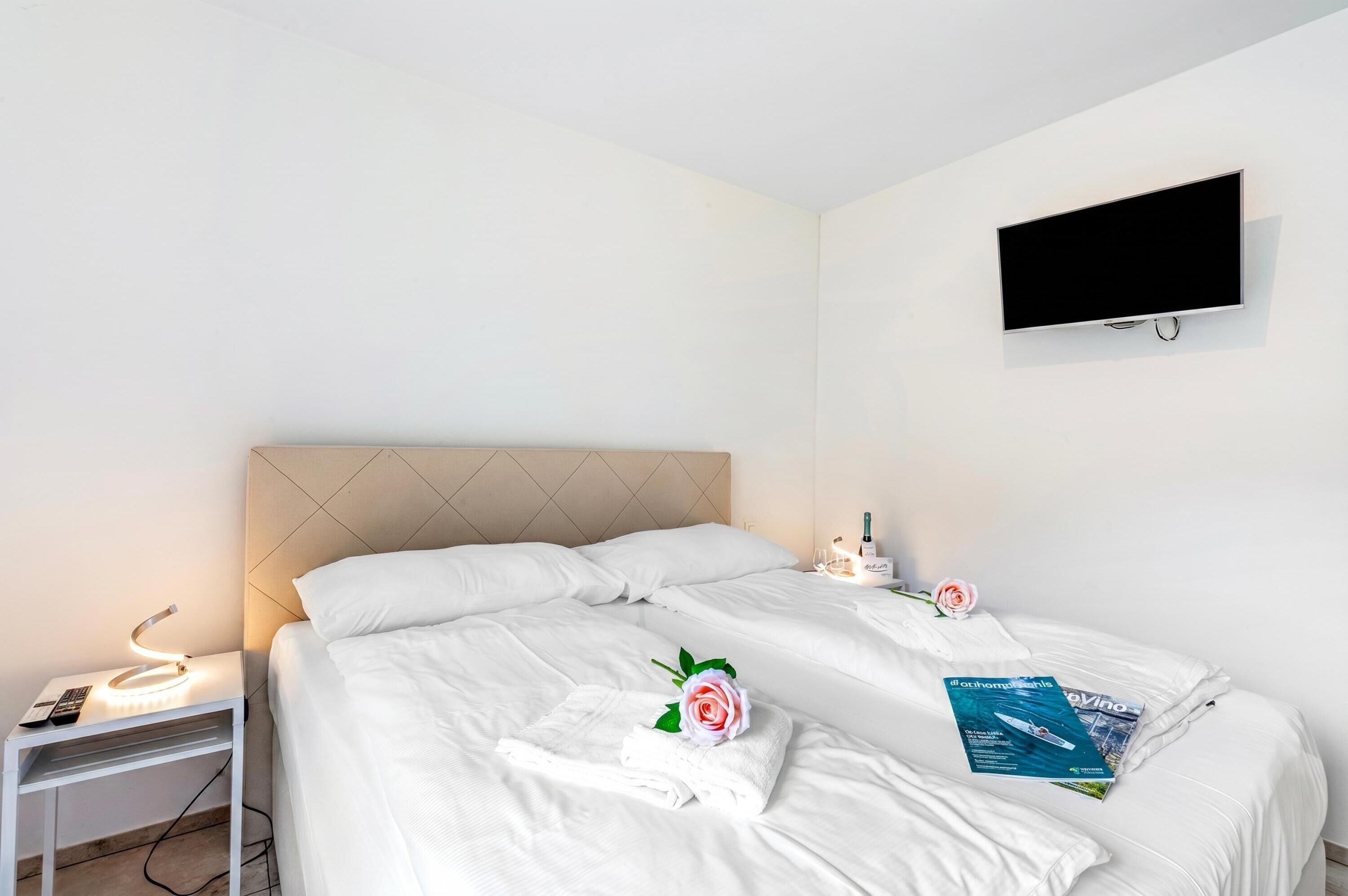 Une chambre d’hôtel avec un lit, une table de chevet avec une lampe, un téléviseur fixé au mur et une rose sur le lit.