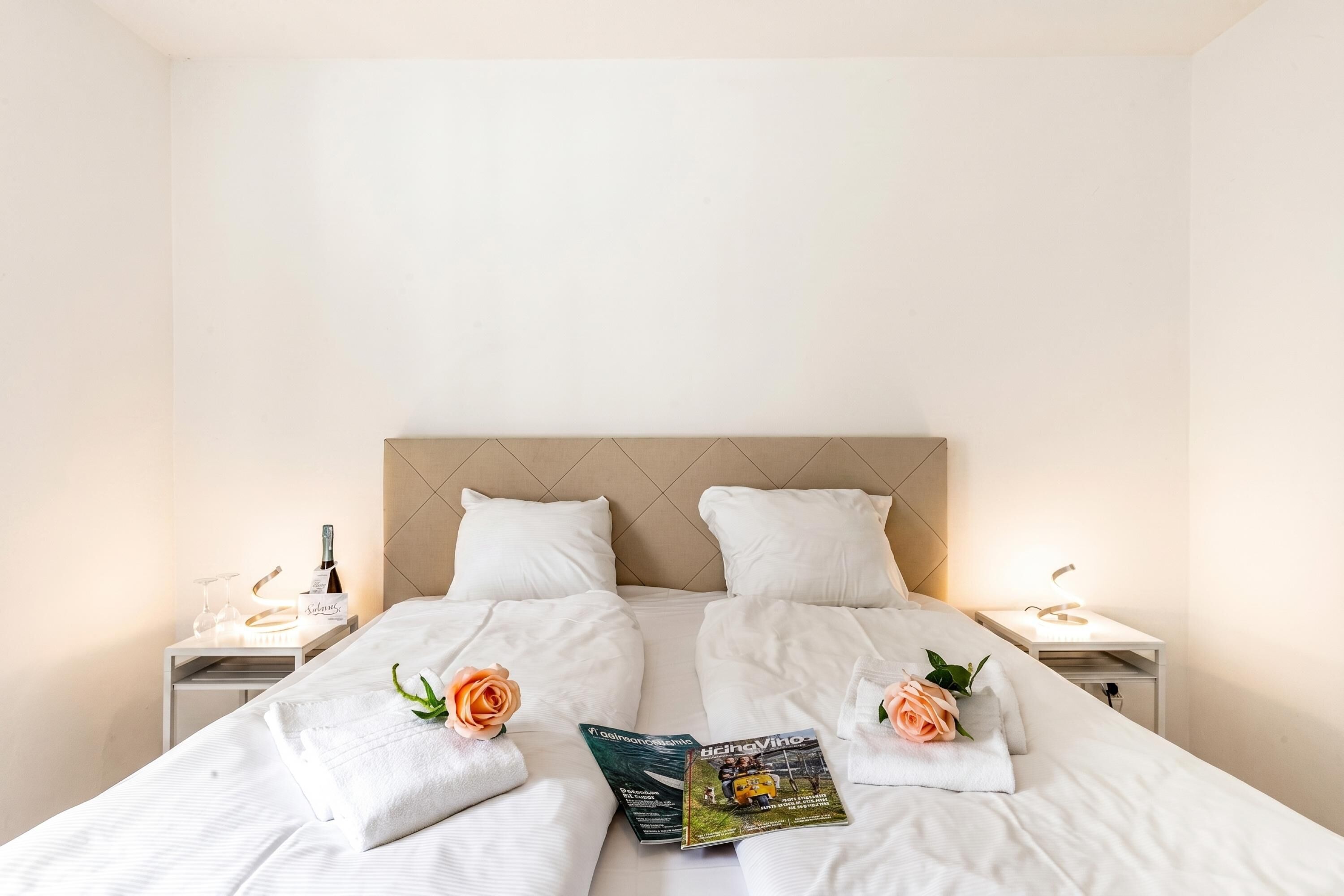 A neatly made bed with white linens, two pillows, a magazine, and a rose on each side.