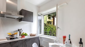 A modern kitchen with a built-in stove, a window with a view of greenery, and a dining area with a table set for two. - Serenata 5 - Cavigliano Studios with Terrace for 5, Cavigliano, Centovalli, Switzerland (Cavigliano, Centovalli)