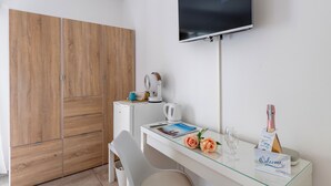Een moderne hotelkamer met een houten garderobe, een wit bureau met een glazen blad, een flatscreen televisie, een kleine witte verwarming, een waterkoker, een fles champagne en een welkomstbord.