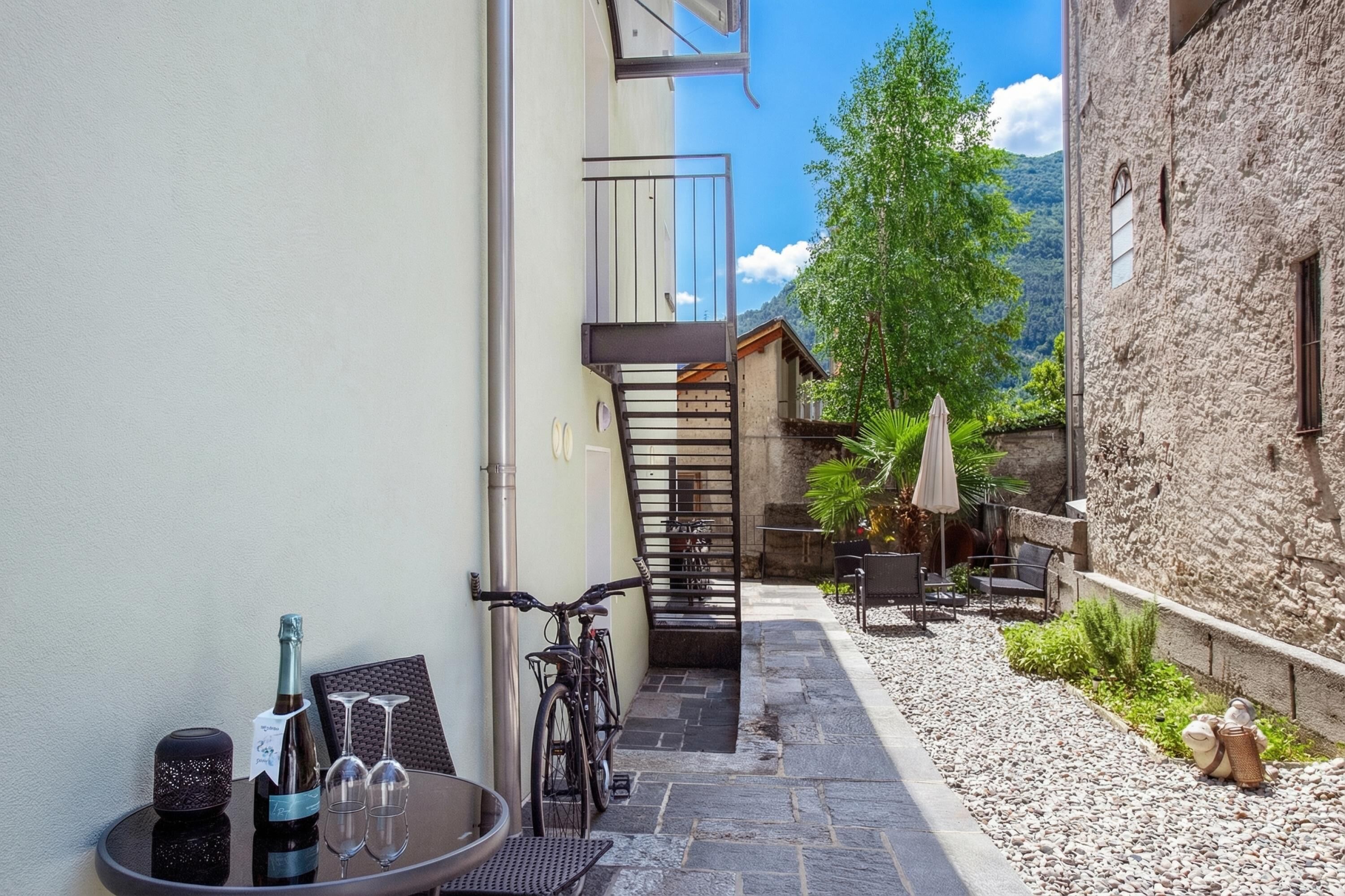 A narrow courtyard with a stone floor, a bicycle, a table with a bottle and glasses, and a chair.
