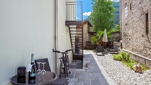 Un estrecho patio con suelo de piedra, una bicicleta, una mesa con una botella y vasos, y una silla.