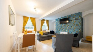 A modern living room with a dining area, a sofa set, a TV mounted on the wall, and a window with yellow curtains. - GuestReady - Charming haven in Knowledge Quarter (Liverpool)