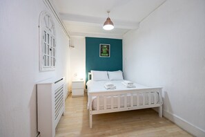 A bedroom with a white bed, a nightstand, a wall-mounted shelf, and a teal accent wall.