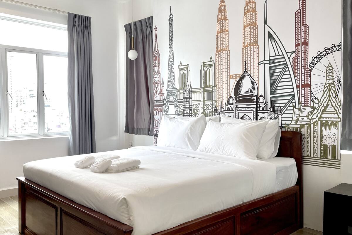 A hotel room with a large bed, grey curtains, a cityscape mural, and a view of buildings through the window.