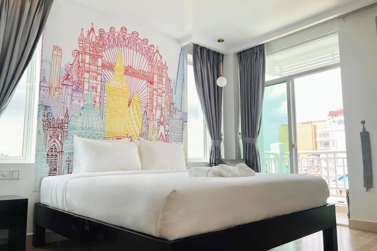A hotel room with a large bed, a cityscape mural on the wall, and a view of buildings through the window.