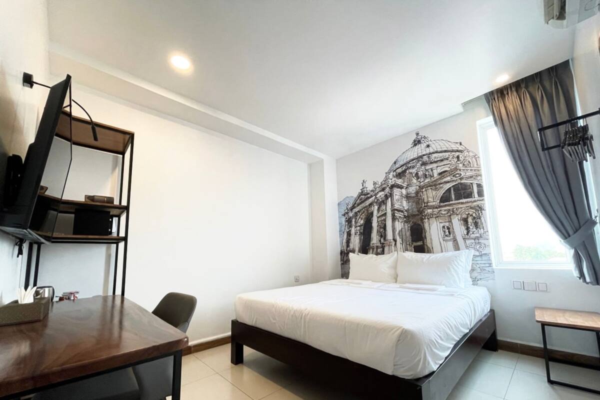 A bedroom with a bed, a desk, and a chair. There is a wall with a mural of a building.