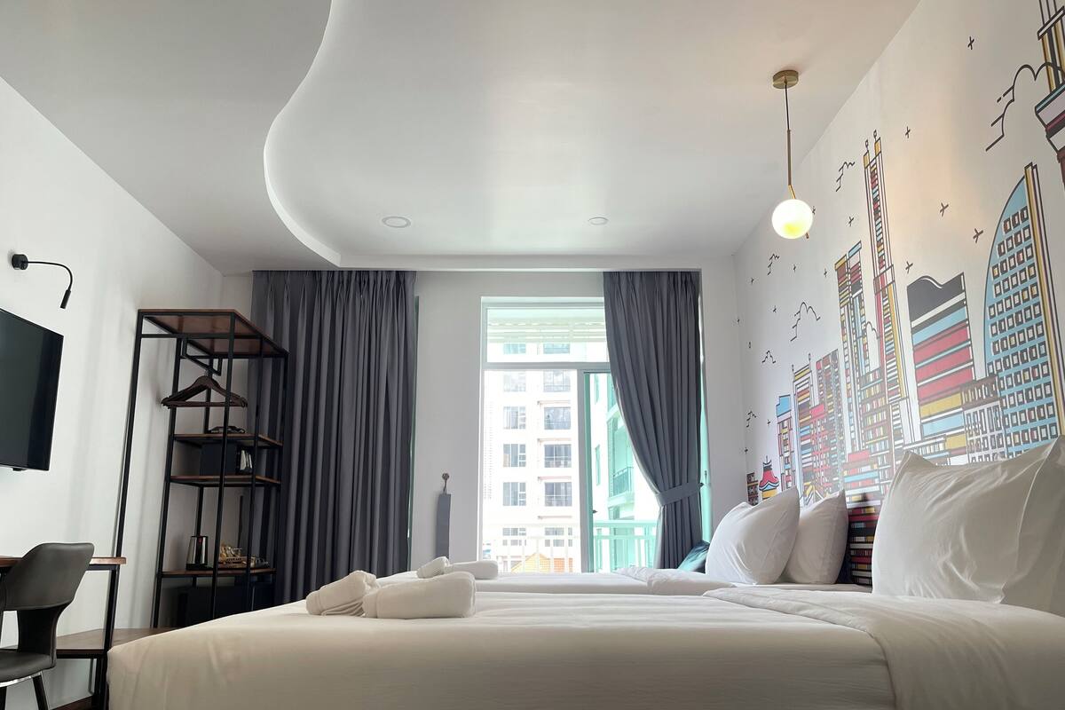 A modern hotel room with a large bed, a desk with a chair, a TV, and a cityscape mural on the wall.