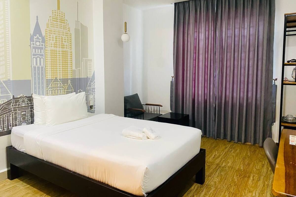 A hotel room with a large bed, a desk, a chair, a bookshelf, and a cityscape mural on the wall.