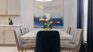 A modern dining area with a white table, grey upholstered chairs, and a blue velvet chair. A painting of flowers and a vase with flowers are on the wall. - Stunning 4bed/3.5 bath pool home, great location, gated community, lake views. (Kissimmee)