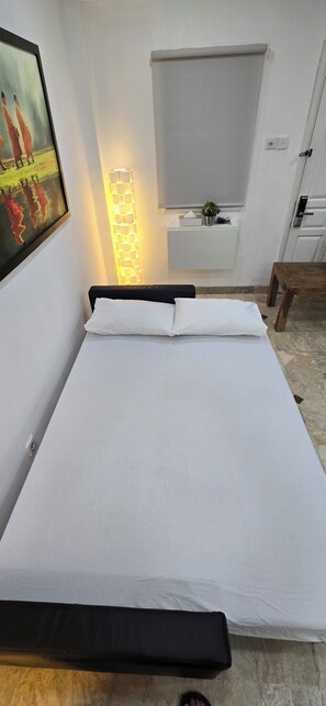 Una cama con sábanas blancas, una mesita de noche, una lámpara de pared y un cuadro en la pared.