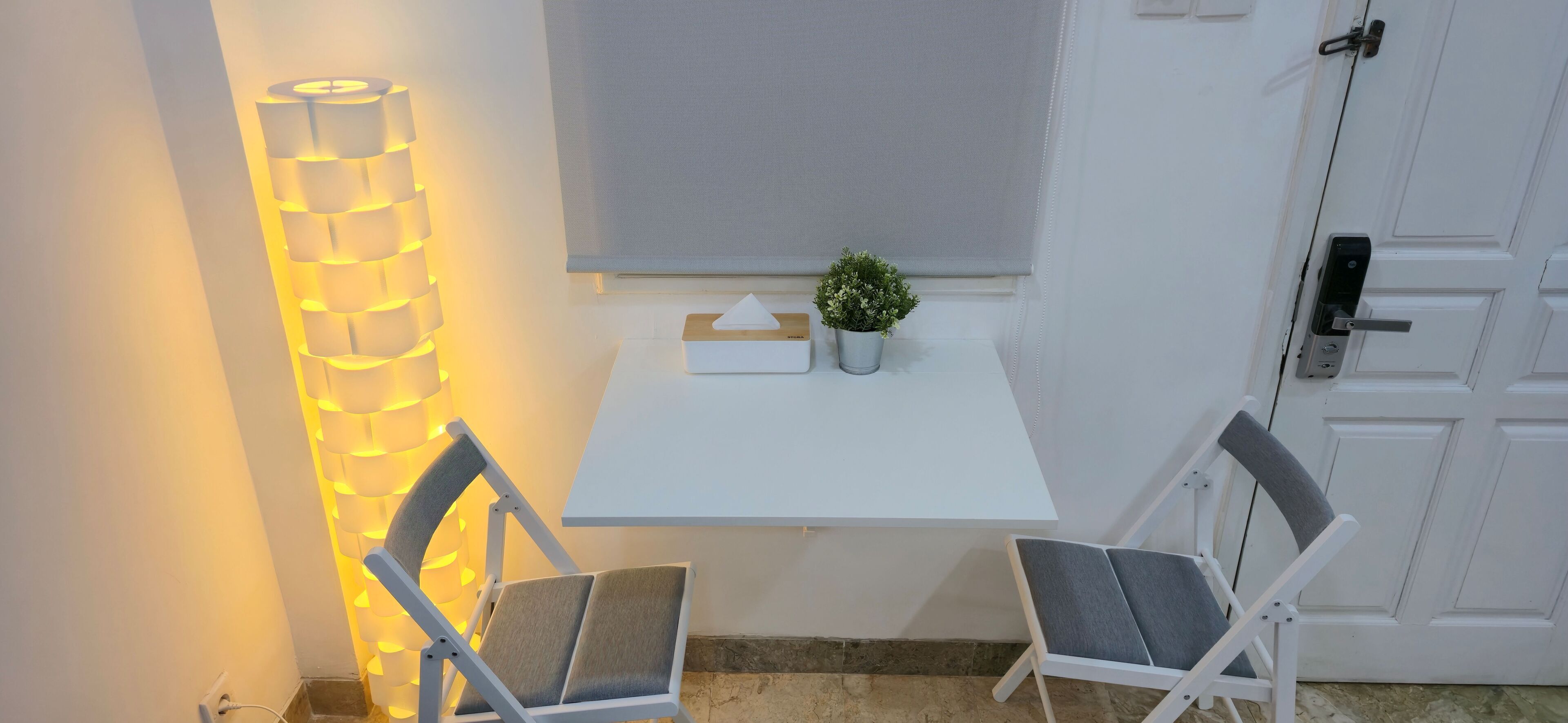 Una habitación pequeña y blanca con una mesa, dos sillas, una planta en maceta, una lámpara de pared y una puerta.