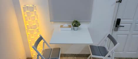 Una habitación pequeña y blanca con una mesa, dos sillas, una planta en maceta, una lámpara de pared y una puerta.
