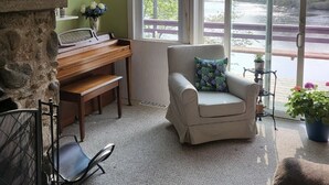 Una habitación acogedora con un piano, un sillón mullido, una chimenea de piedra y vistas a un río a través de amplias ventanas.