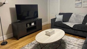 A living room with a dark-colored sofa, a flat-screen TV on a black stand, a round coffee table, and a grey rug.
