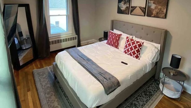 A bedroom with a large bed, a bedside table, a nightstand, a window with curtains, a mirror, and a wall with three framed pictures.