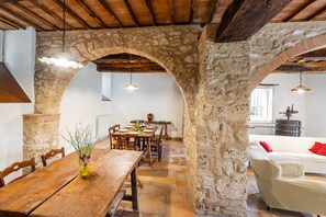 Un comedor rústico con una mesa y sillas de madera, arcos de piedra y un sofá blanco en un ambiente acogedor.